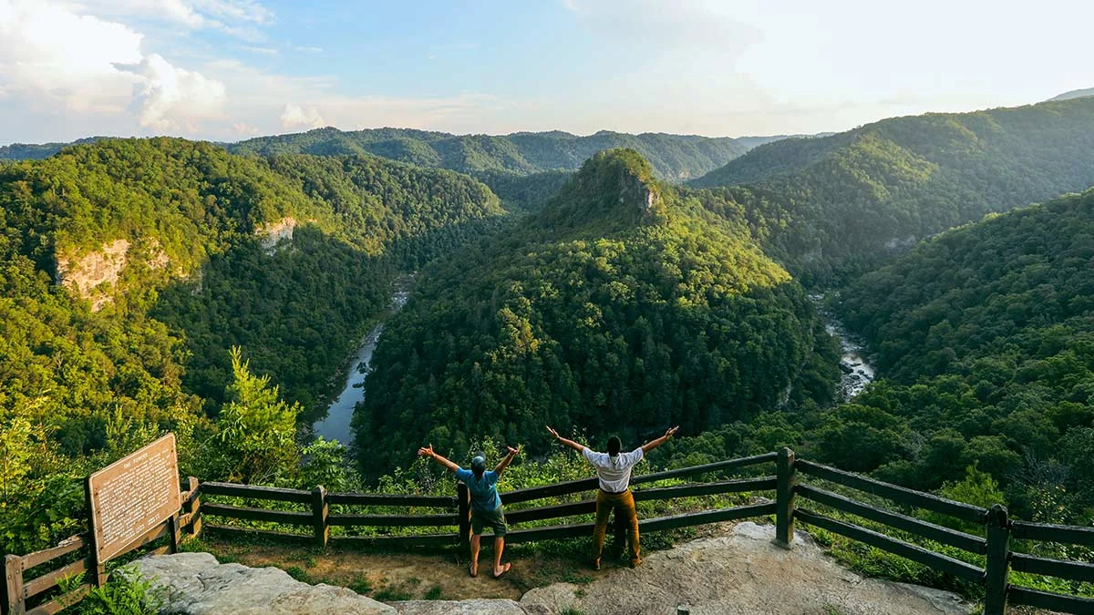 Two people with arms raised at a Breaks Interstate Park overlook above the forested river gorge