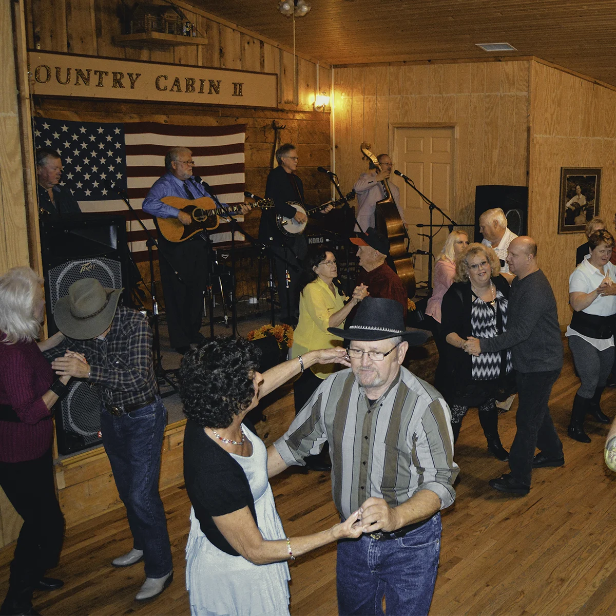 A bluegrass band with guitar, banjo, and upright bass performs beneath an American flag while couples dance on the wooden floor at Country Cabin II in Norton, Virginia.