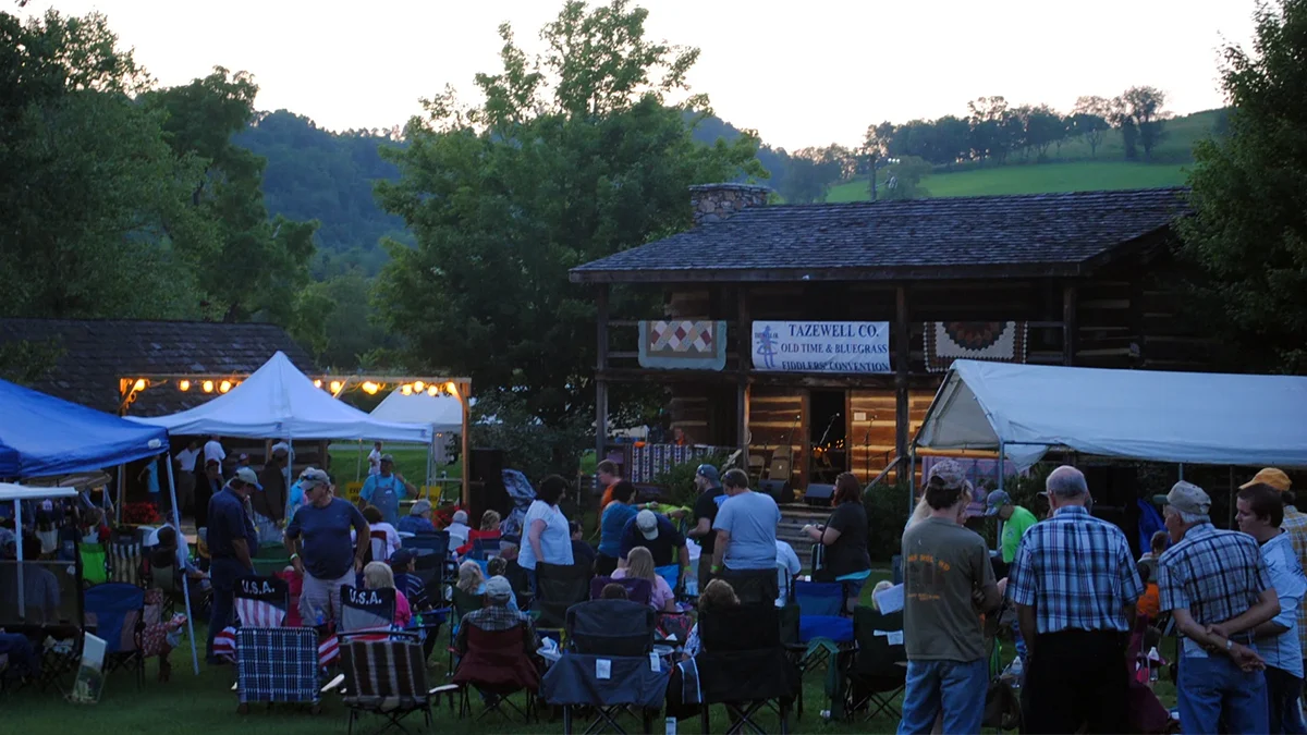 A crowd gathers at dusk before a log cabin stage for the Tazewell County Old Time and Bluegrass Fiddlers Convention in Tazewell, Virginia.