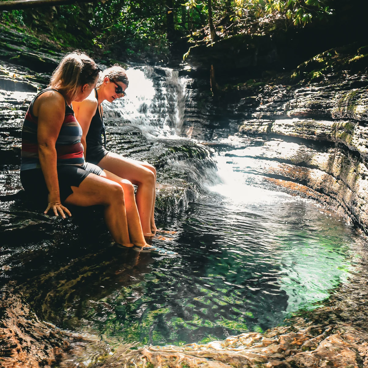 Two visitors sitting at the edge of the emerald Devil's Bathtub swimming hole below a small waterfall in Scott County, Virginia