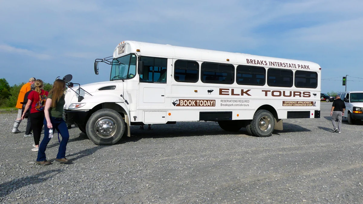 Visitors walking up to board the white Breaks Interstate Park Elk Tours bus in a gravel parking lot in Southwest Virginia.
