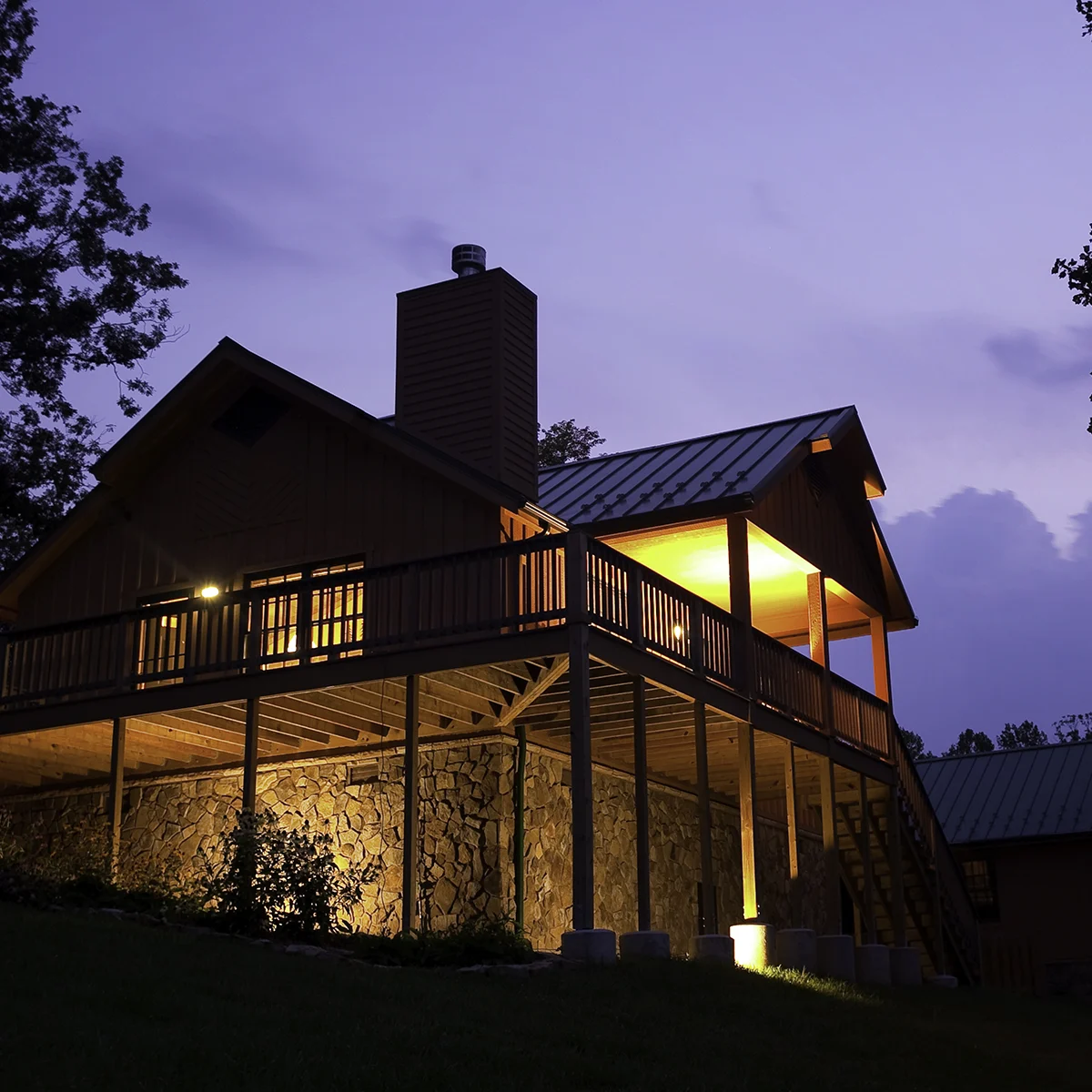Elevated cabin with lit wraparound deck and stone foundation glowing at dusk in the Southwest Virginia mountains.