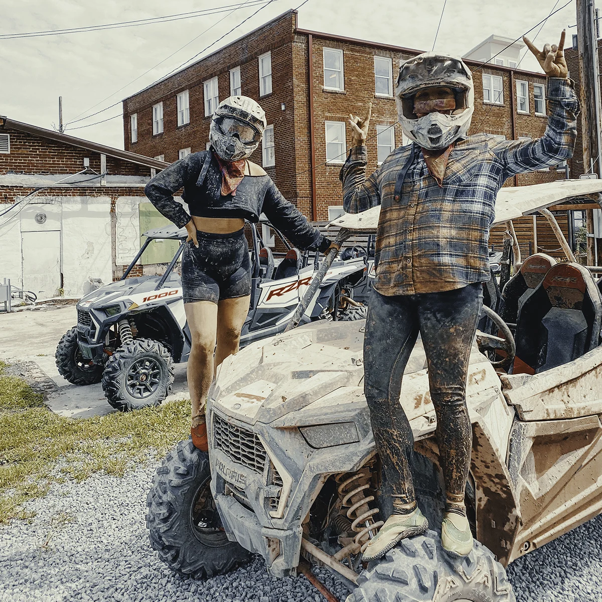 muddy-utv-riders-polaris-rzr-town-virginia.jpg Two mud-covered off-road riders wearing full-face helmets pose on and beside their muddy Polaris RZR UTVs in front of a brick building in a Virginia trail town.