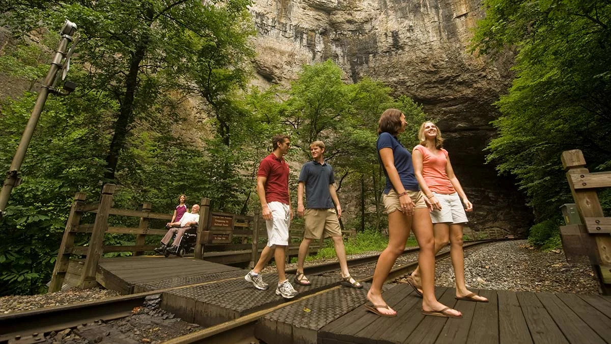 Group walking the boardwalk toward the cave entrance at Natural Tunnel State Park, with a wheelchair user on the path