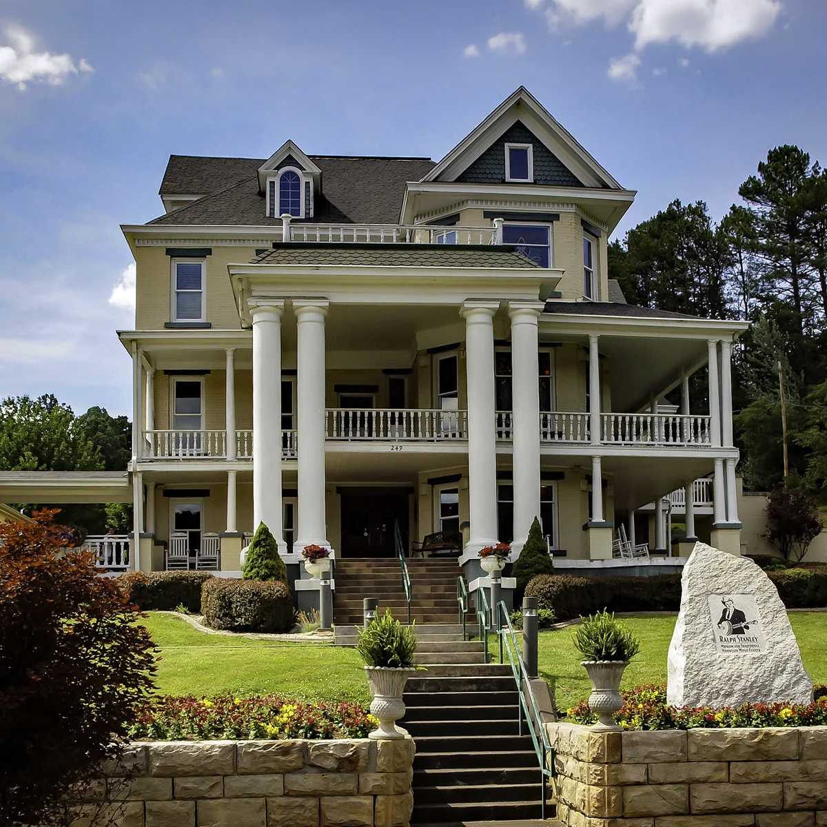 Exterior of the Ralph Stanley Museum and Traditional Mountain Music Center, a restored yellow Victorian house with white columns and double wraparound porches, with a Ralph Stanley stone marker in the foreground.