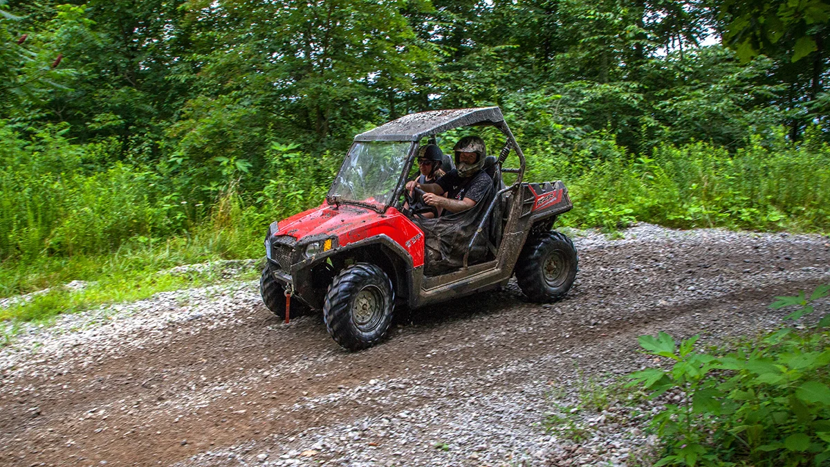 red-polaris-rzr-side-by-side-wooded-trail-virginia.jpg A mud-splattered red Polaris RZR UTV driving through a dense, green forested off-road trail in Virginia.