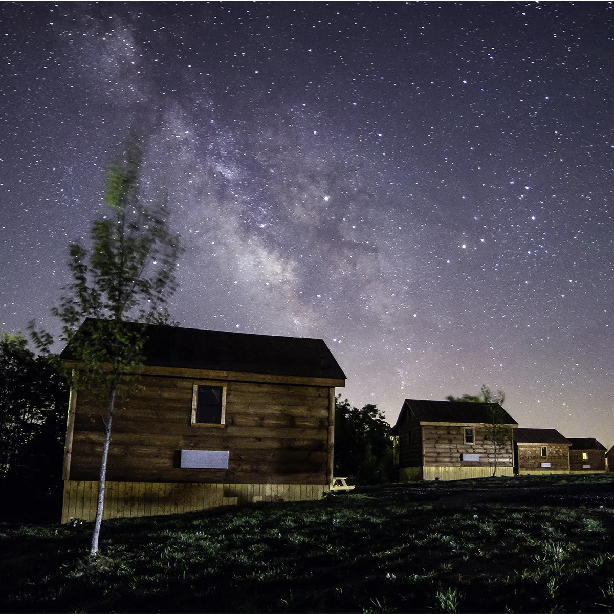 Row of rustic log cabins under a star-filled sky with the Milky Way visible, Southern Gap Outdoor Adventures lodging at night.