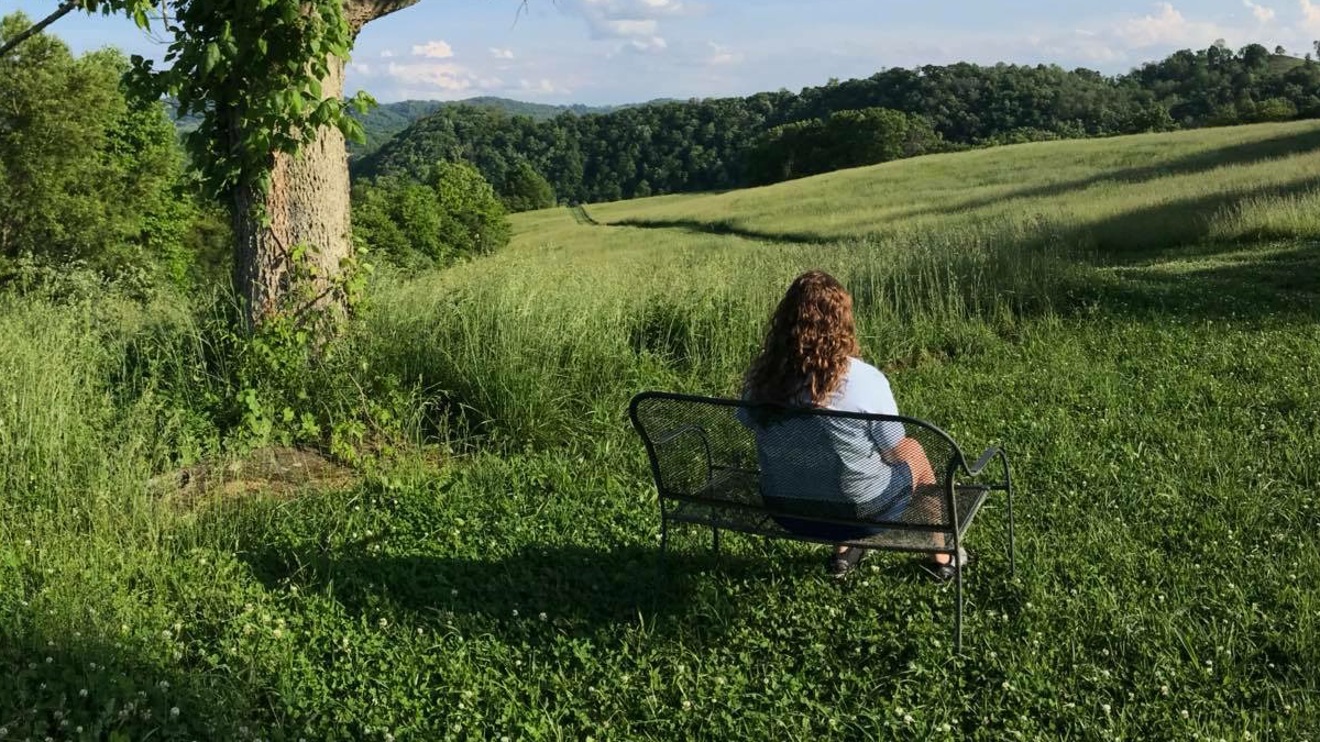 A visitor resting on a bench overlooking a rolling green meadow and forested ridges along the Sugar Hill Loop in St. Paul, Wise County.